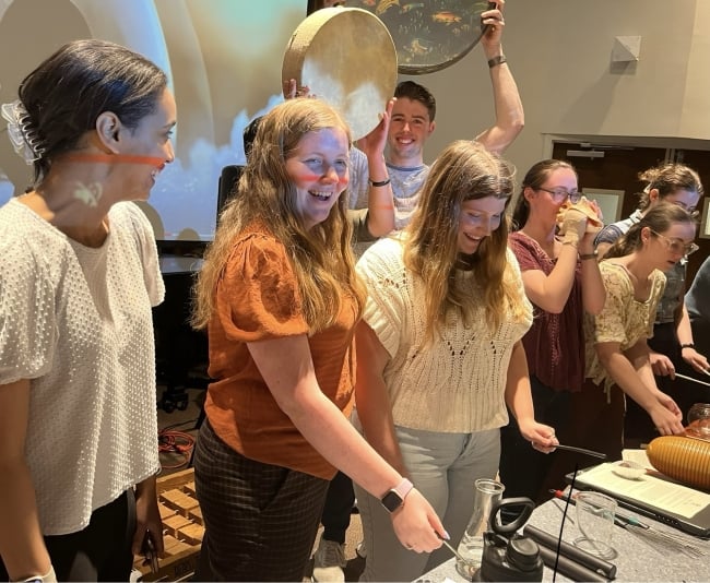 A group of students hold and play musical instruments in a classroom, smiling.