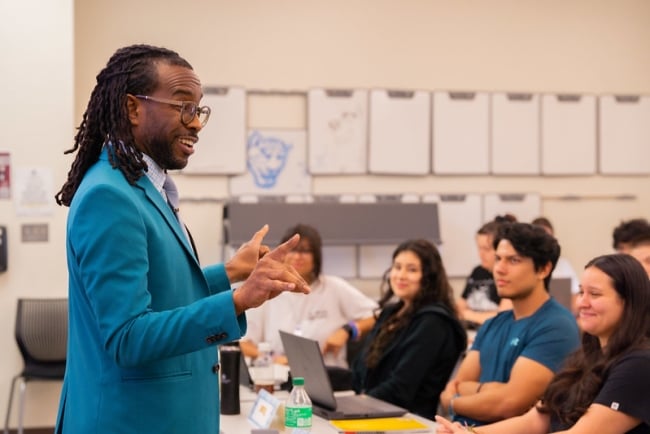 A dark-skinned man with shoulder-length hair wearing a blue jacket shares a story with students in his class, some of whom are smiling.