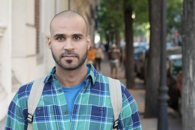 A man with a short haircut, goatee and plaid shirt, wearing a backpack, walks on a city sidewalk.