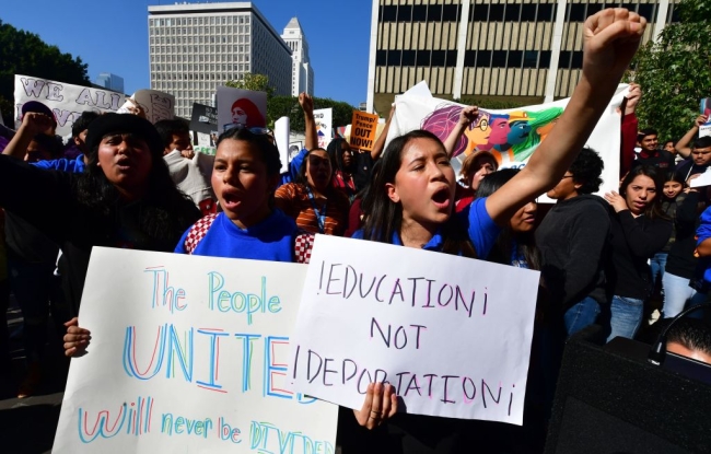 Two young people yell while holding signs, one raising a fist. A sign reads "education not deportation."