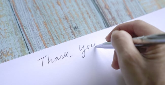A close-up image of the hand of a person holding a pen as they write "Thank you" on a blank note card.