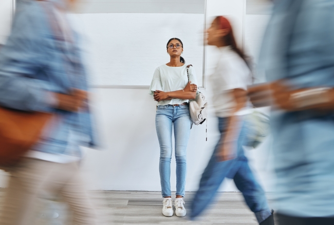 Young woman stands still in a hallway looking pensive as other young people rush by.