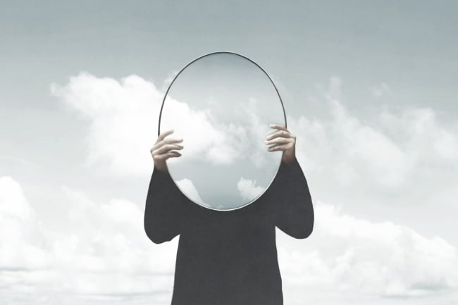 An illustration of a woman holding an oval mirror up to her face against a cloudy gray-sky background.