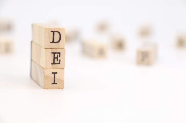 A vertical stack of three wooden blocks with letters on them reads "DEI." Other blocks, their letters blurry, can be seen strewed in the background.