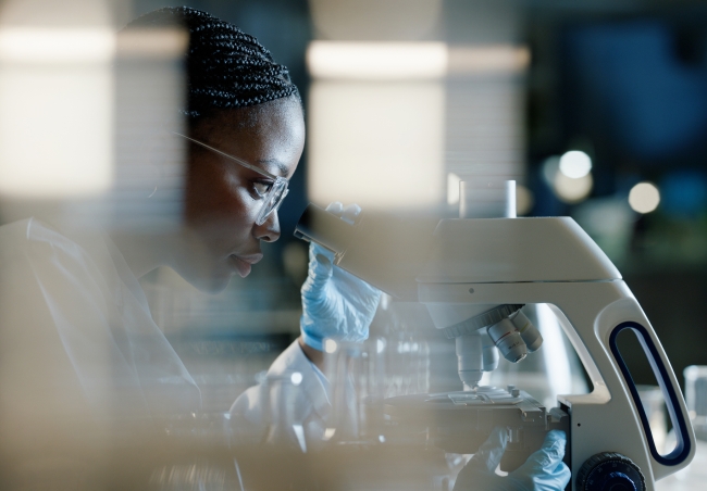 A scientist wearing protective eyewear and gloves looks into a microscope in a lab.