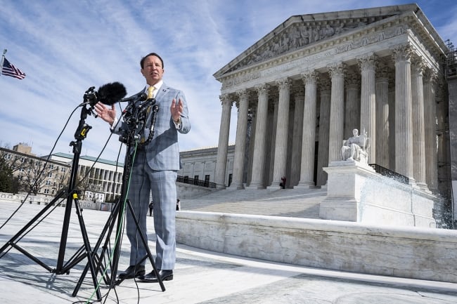 A photograph of Louisiana governor Jeff Landry standing in front of a couple of microphones outside the U.S. Supreme Court building.