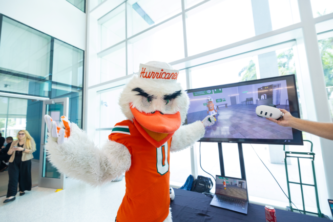 The University of Miami mascot holds VR controllers in front of a TV display