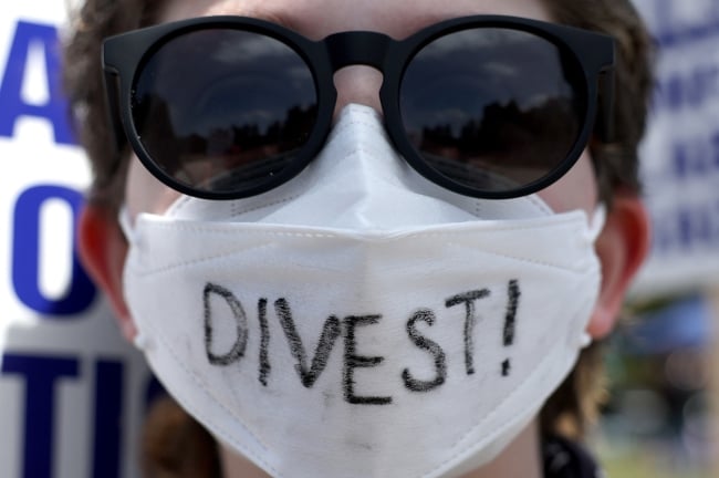A photograph of the face of a person wearing sunglasses and a white face mask, on which "Divest!" is written.
