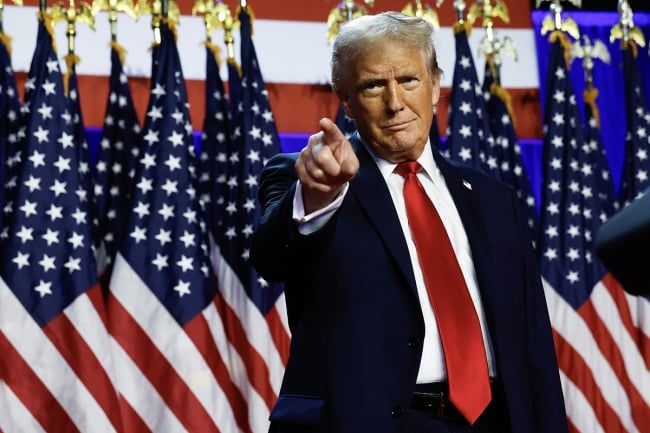 Donald Trump, in a suit and red tie, stands in front of a row of American flags and points his finger.