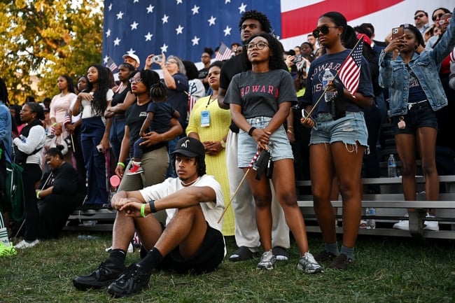 People sit and stand on a patch of grass in front of an American flag