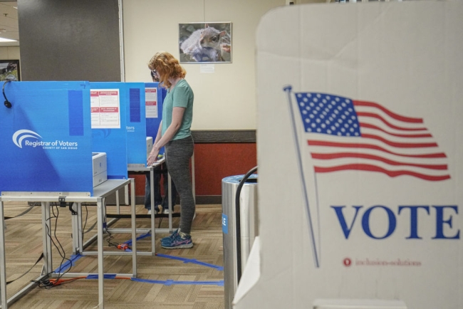 In the foreground, the image of an American flag with the word "vote" below it. In the background, a person with blond hair is seen filling out a ballot.