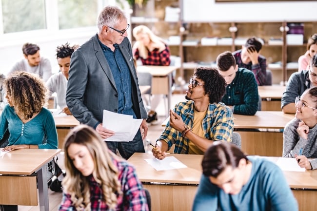 A professor speaks to an engaged student in a classroom full of students.