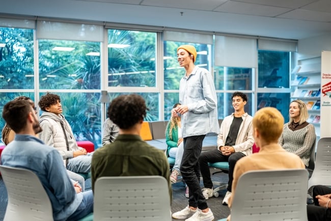 Young man in casual clothes stands in the middle of a seated circle of peers and a facilitator. 