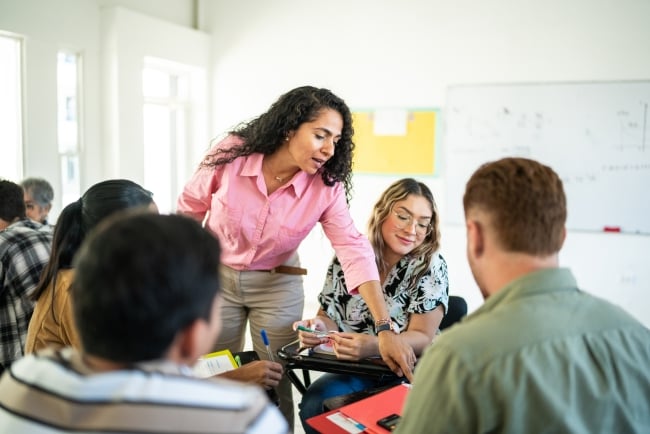 A professor talks and points to a notebook on student's desk while students sit in a circle.