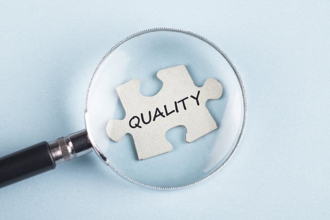 An illustration of a jigsaw puzzle piece with the word "Quality" held under a magnifying glass.