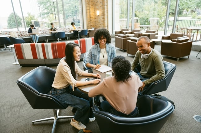 In a student center lounge space, four students sit around a table as if running a meeting.