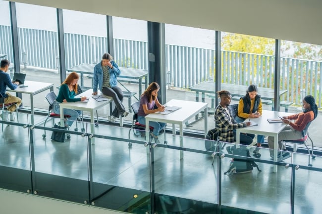 University students are seen scattered at various desks in an open study area, as they work on assignments between classes. Some students are working together while others are seen working independently in this aerial view.