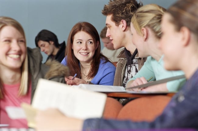 In the forefront of the picture, group of five students are gathered in a lecture hall, talking and smiling with one another.