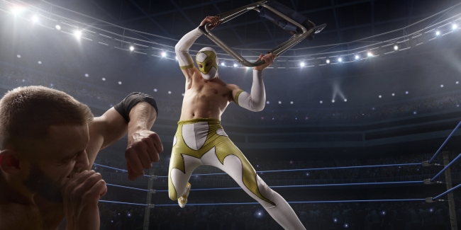An image of two professional wrestlers in a match. One lunges over the other while the other seemingly cowers, protecting his head with his arm from a coming blow.