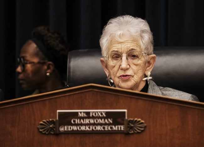 An older light-skinned woman with white hair, wearing glasses, at lectern