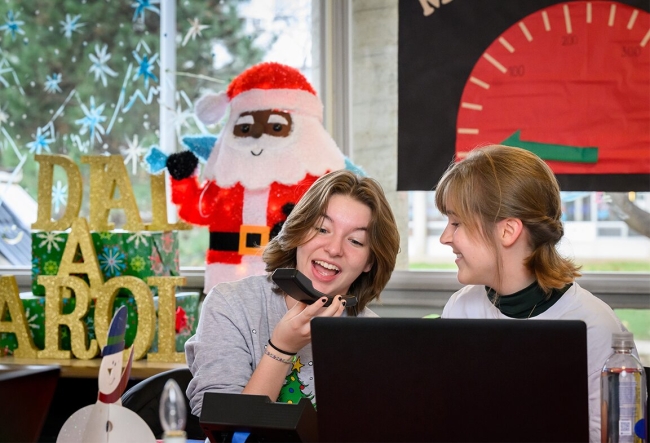 Two students answer the phone singing in a Christmas-decorated room
