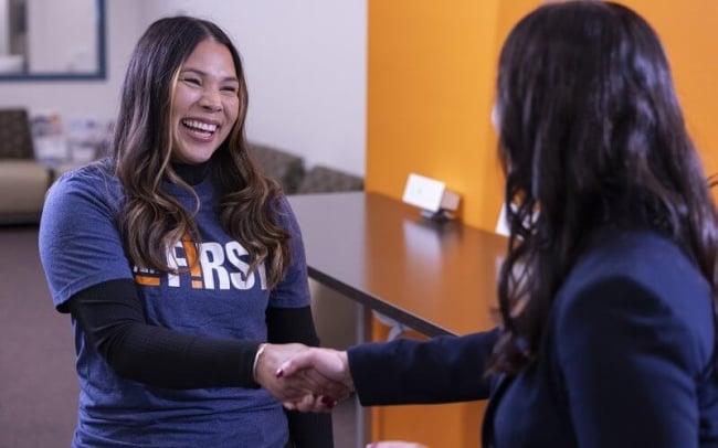 A young woman shakes another person's hand while smiling, wearing a sweatshirt that says I Am First