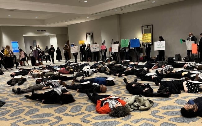 A photograph of a "die-in" protest at the Modern Language Association annual convention. Dozens of people are lying on the floor in a hotel, and people are standing around them with signs.