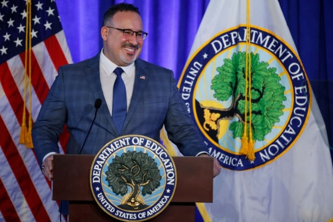 Miguel Cardona stands a podium in front of Education Department and American flags