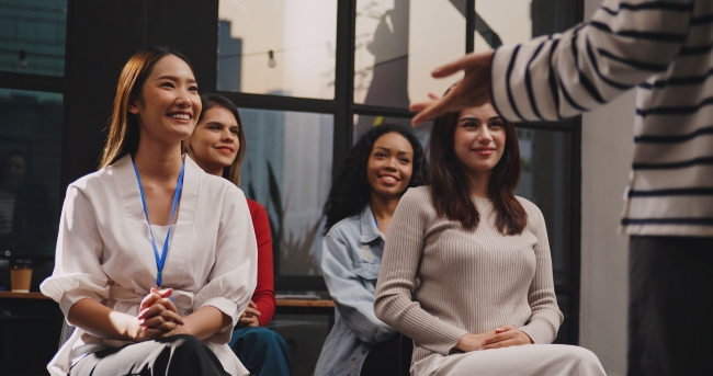 A group of four young women smile and listen attentively in the audience for a lecturer or seminar presenter.