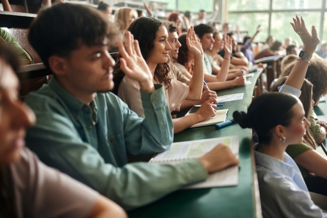 Large group of university students raising their hands to ask the question on a class in amphitheater. Focus is on smiling woman.