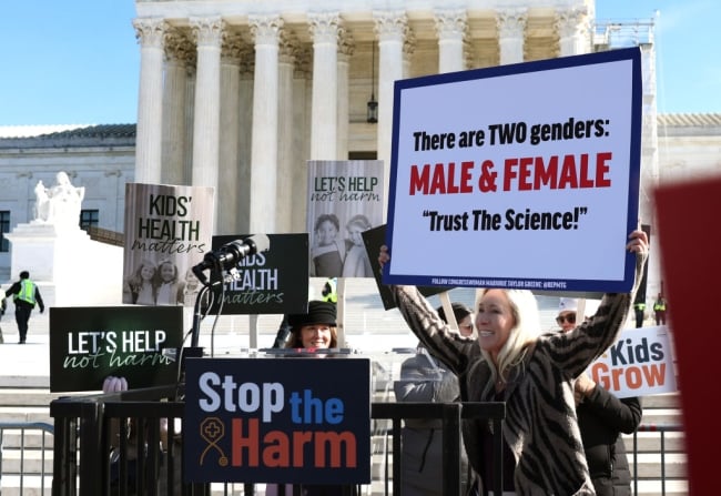 A blonde congresswoman holds up a sign that says there are "two genders, male and female," and "trust the science."
