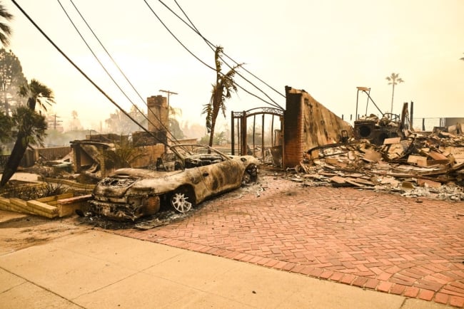 A burned out car sits in front of the ruins of a house.