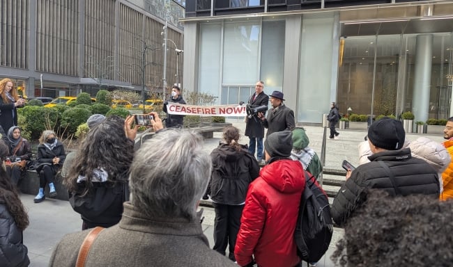 A photograph of a man speaking into a megaphone at a rally next to two people holding a banner reading "Ceasefire Now!"