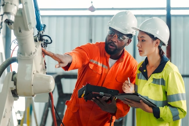 An instructor in a hard hat instructs a student in a hard hat, pointing to a machine.