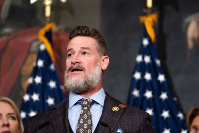 Greg Steube, a white man with a beard, is pictured in front of two American flags.
