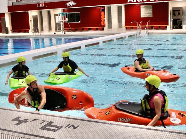 Five students sit in kayaks in an indoor swimming facility