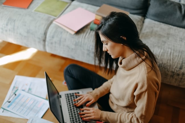 A young woman types on a laptop with papers strewn around her.