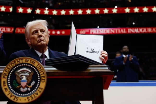 Donald Trump sits at a desk with the presidential seal, holding up an executive order he signed.