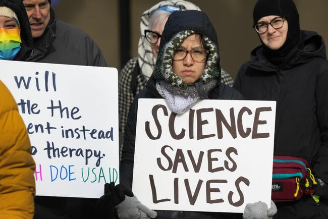 A person in a jacket holds a white sign that says "science saves lives" in black text