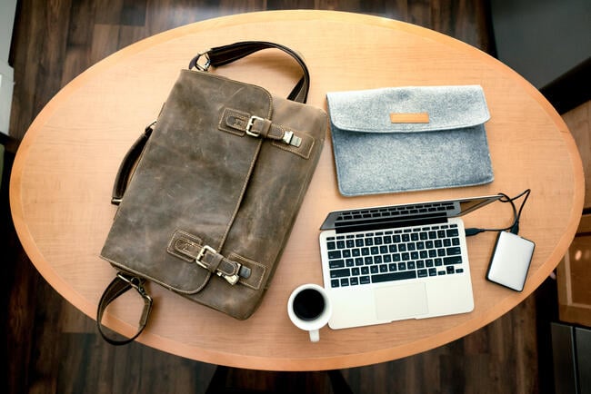 A photo taken from overhead of a shoulder bag, a laptop sleeve, a laptop, an external hard drive and a cup filled with coffee, all atop an oval-shaped table.