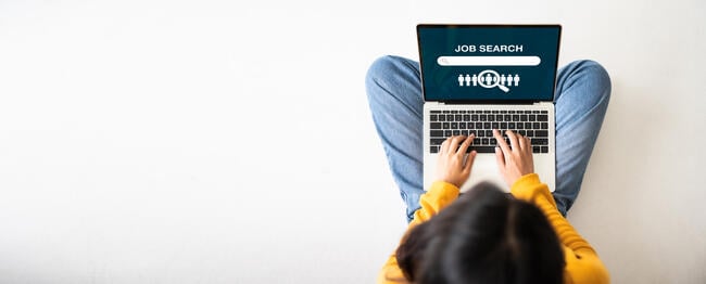 A young adult woman dressed casually in jeans and a sweater sits cross-legged on the floor, with a laptop open to a page that says "Job Search." The photo is taken from overhead.