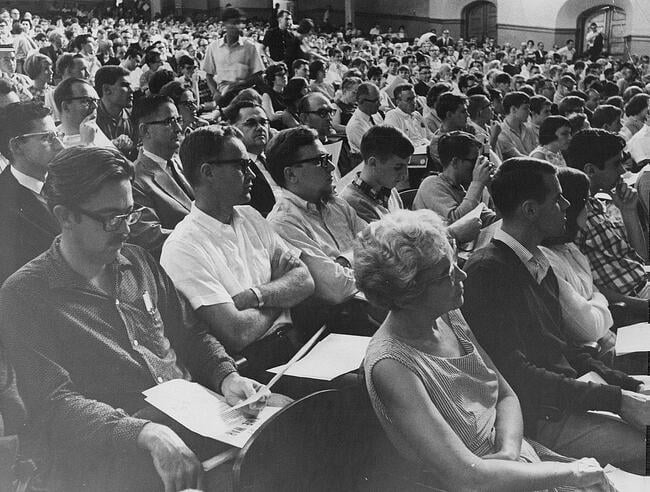In this historical black-and-white photo from a 1965 anti-war teach-in, a dense crowd of people in an auditorium sit in an audience, seemingly listening to a speaker with intense interest and focus.