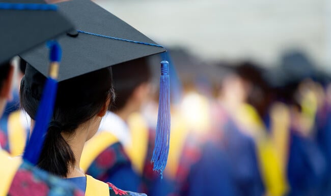 A rear view of graduates during commencement wearing blue and yellow regalia and graduation caps.