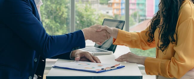 Two people shaking hands in front of a laptop. The laptop is open and has a picture of a person on it.