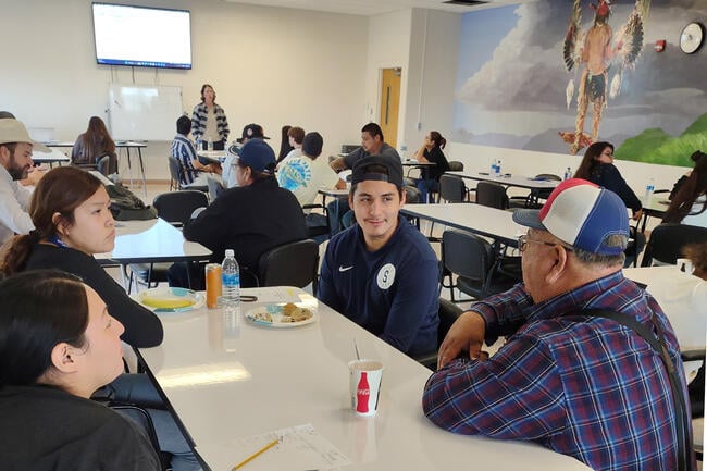 Students sit around a table talking with a Native American mural in the background.