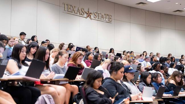 A group of students sit in a large lecture hall, facing the front of the room. Behind them on a large white wall is the Texas State logo.