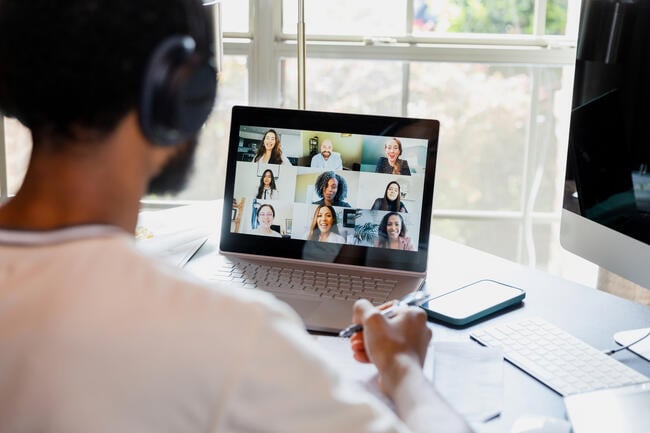 Young man looks at a computer screen with friendly-looking classmates in a grid. He is wearing headphones and faced away from the camera.