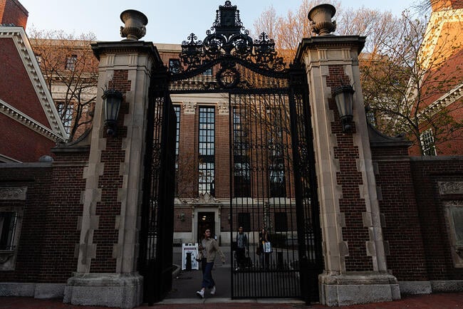 A photo of a student walking through campus gates at Harvard University, a stately brick campus building in the background.