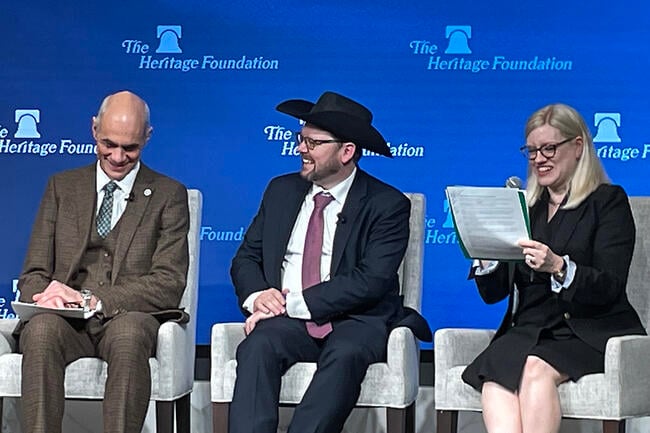A photo of three panelists speaking at the Heritage Foundation. The man in the center is wearing a cowboy hat.