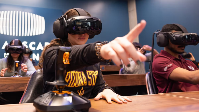 A classroom of students wearing black VR goggles, the student in the foreground has one hand outstretched in front of them